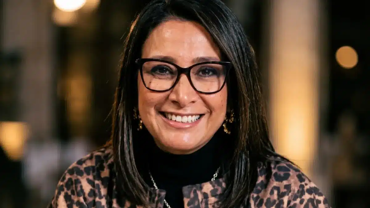 Un primer plano de Claudia Benavides Salazar, Ministra de Ciencia, Tecnología e Innovación de Colombia, sonriendo cálidamente. Lleva gafas y una chaqueta con un patrón texturizado marrón y negro. El fondo está desenfocado.
